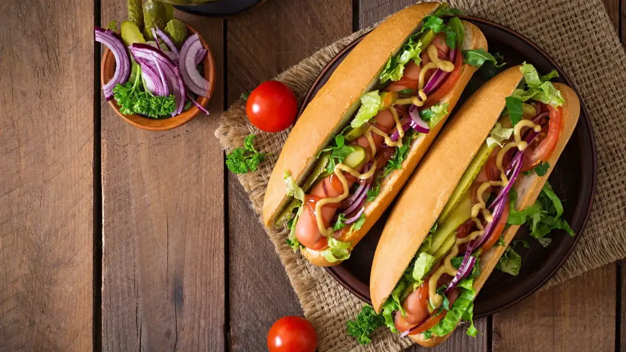 Dwa apetyczne hot dogi z bułką, parówką, sałatą, pomidorem, ogórkiem, czerwoną cebulą i musztardą. Idealny hot dog przepis na szybki obiad.