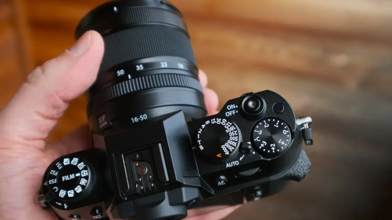 Dłoń trzyma aparat Fujifilm XT50 z obiektywem 16-50mm. Widoczne pokrętła tryb&oacute;w filmowych i ekspozycji.