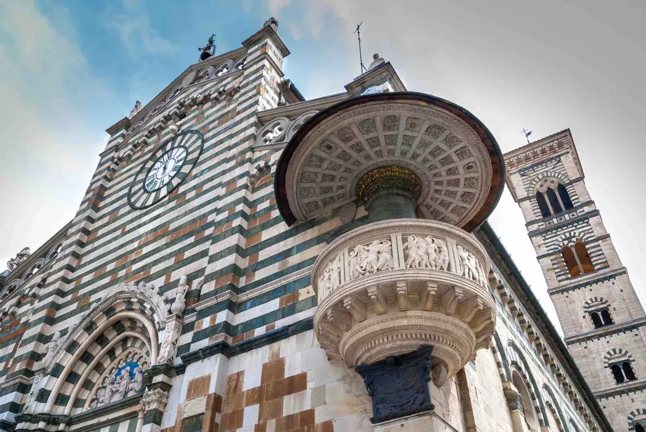 Il Duomo di Prato, con la sua facciata a strisce bianche e verdi, domina il prato della città. Un pulpito decorato con bassorilievi e un campanile svettano nel cielo azzurro.