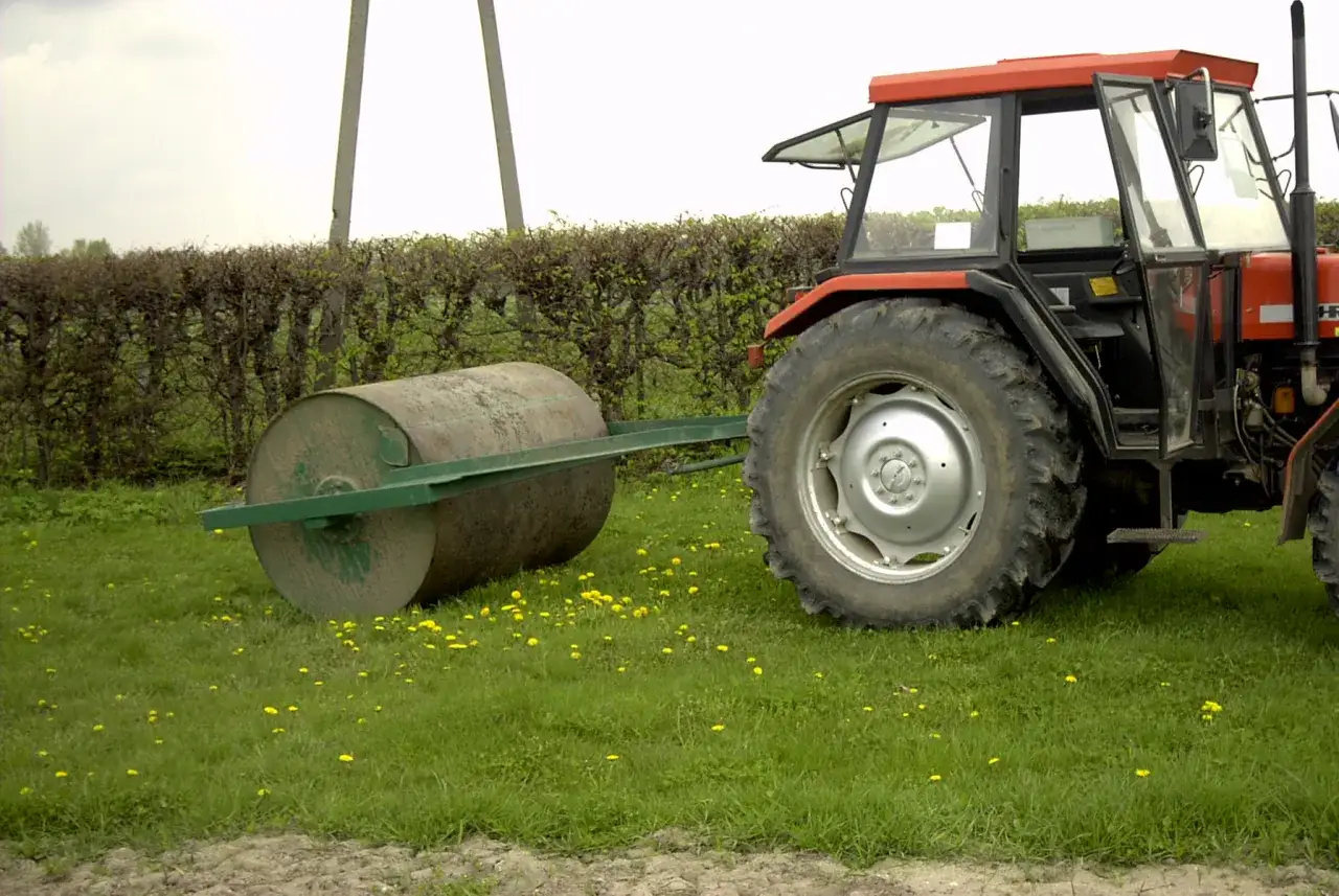 Czerwony traktor z przyczepionym walcem czeka na rozpoczęcie wałowania trawnika.