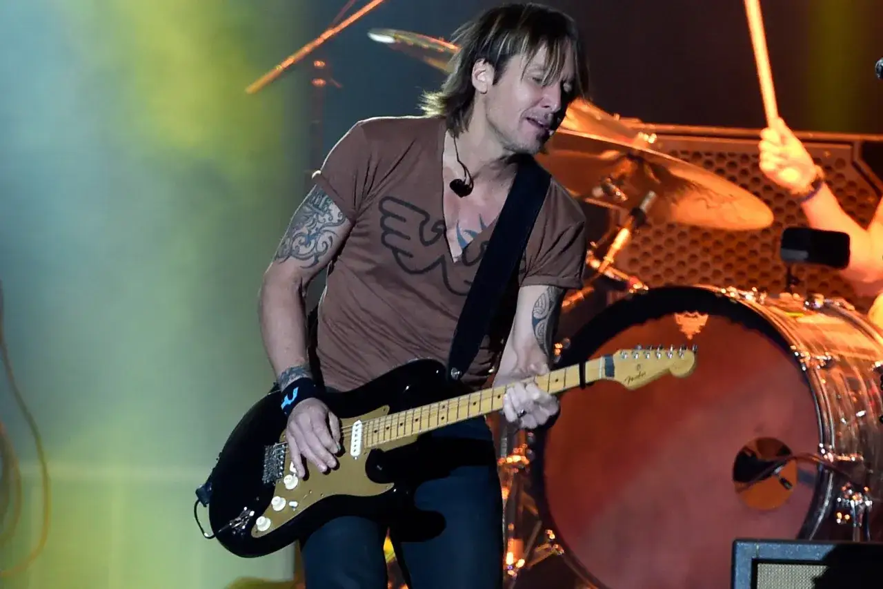 Keith Urban gra na gitarze, a za nim perkusista przygotowuje się do występu. To świetny przykład energii, jaką niosą ze sobą zespoły rockowe.