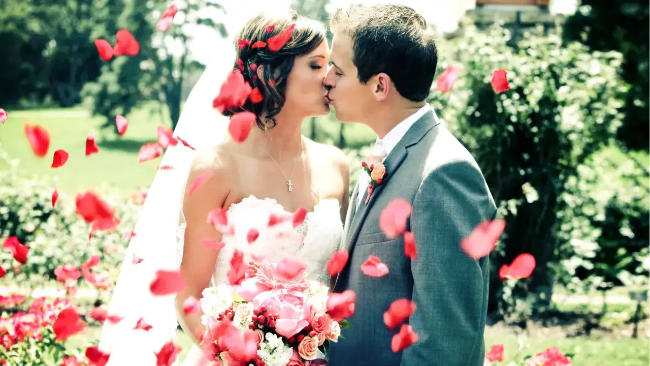 Una pareja se besa rodeada de pétalos rojos, como si fuera un sueño de boda.