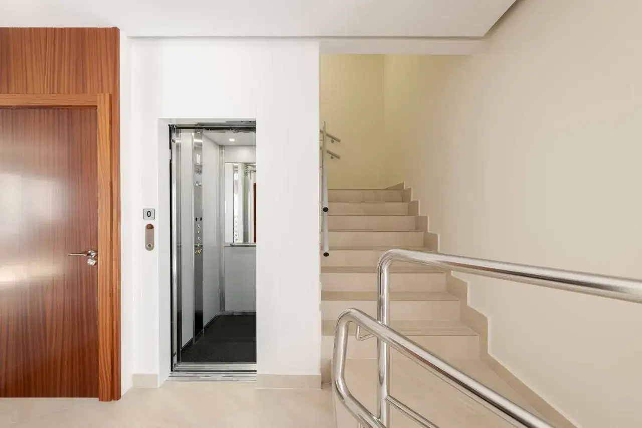 Puerta de madera, ascensor abierto y escaleras con barandilla met&aacute;lica, elementos comunes de un edificio moderno.