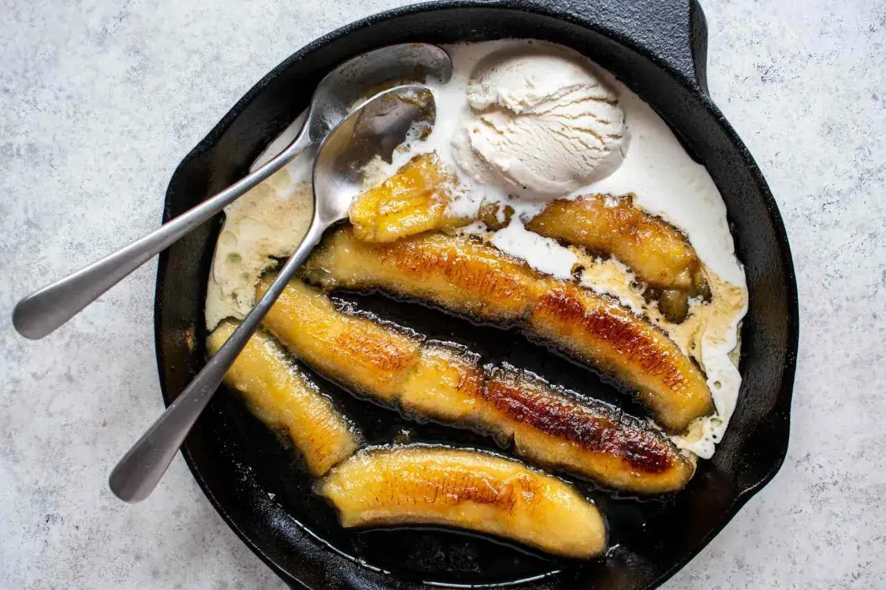 Bananes flamb&eacute;es caram&eacute;lis&eacute;es dans une po&ecirc;le en fonte, servies avec une boule de glace &agrave; la vanille et deux cuill&egrave;res.