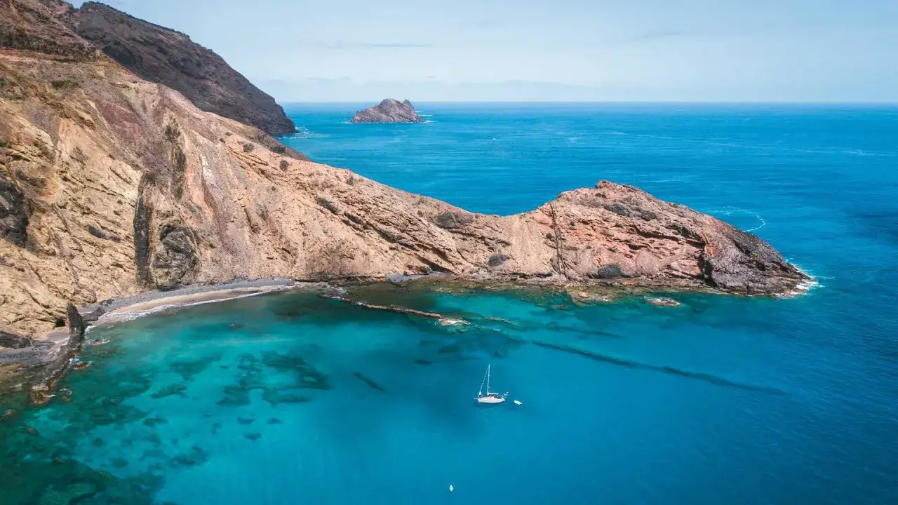 A lone sailboat drifts in the turquoise waters off the rugged coast of Porto Santo, with dramatic cliffs meeting the sea.