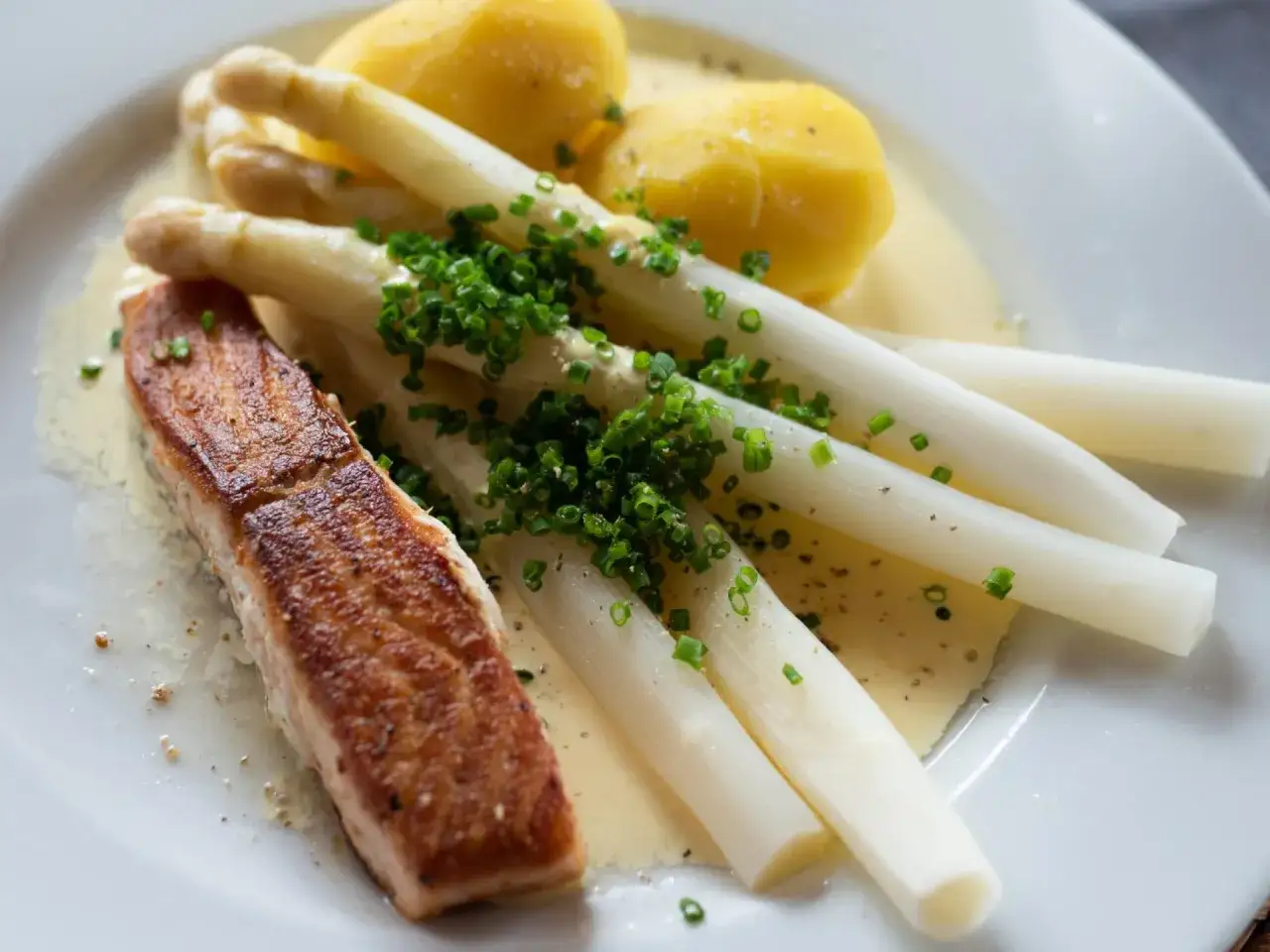 Ein Stück gebratener Lachs liegt neben einem Bund weißem Spargel, garniert mit Schnittlauch. Dazu gibt es Salzkartoffeln und Sauce Hollandaise. Ein Genuss, der den Spargel stillen lässt.