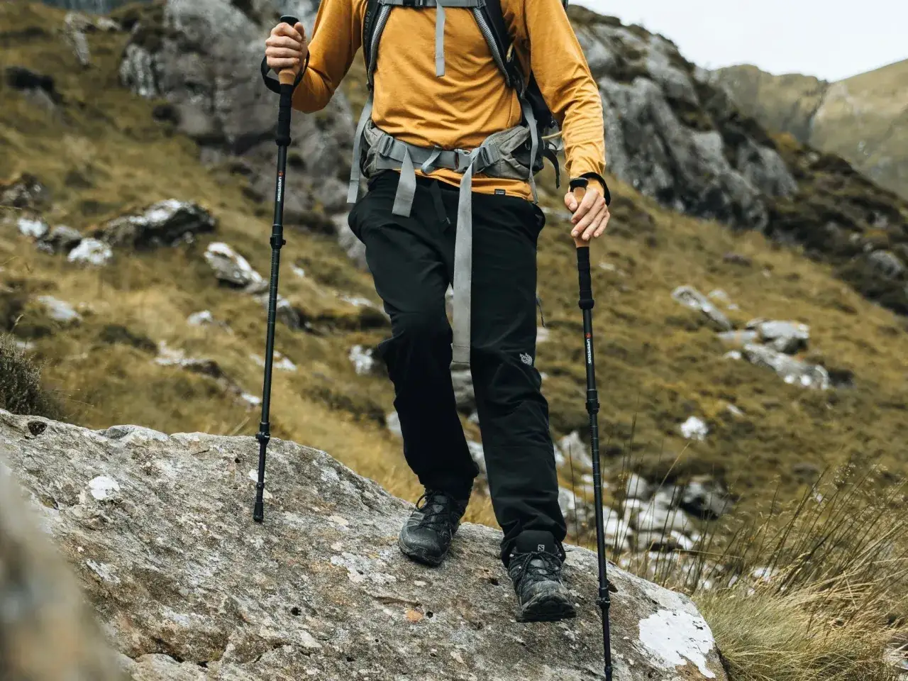 Osoba w ż&oacute;łtej bluzie i czarnych spodniach z kijkami trekkingowymi pokonuje kamienisty szlak. To świetny przykład, jak chodzić z kijkami trekkingowymi.