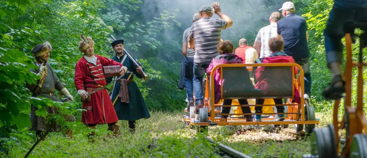 Bieszczady z wózkiem. Grupa osób w historycznych strojach i turyści na drezynach przemierzają leśną ścieżkę.