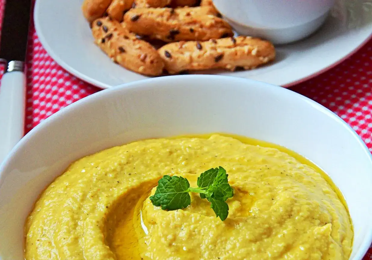 Gładki, żółty hummus z fava fasoli, ozdobiony miętą, podany z chrupiącymi paluszkami.