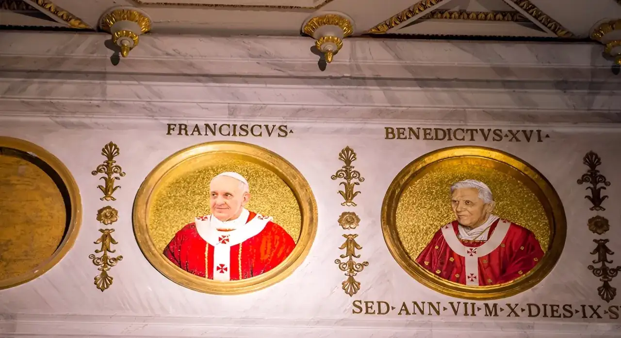 Portrety papieży Franciszka i Benedykta XVI. Złote medaliony z imionami papieży zdobią marmurową ścianę.