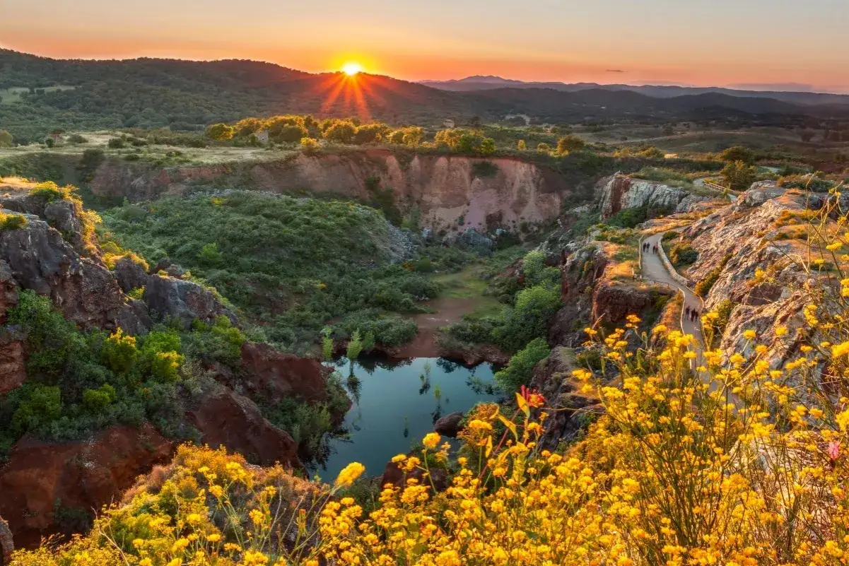 Atardecer en las rutas senderismo Sevilla. Un camino serpentea entre rocas y flores amarillas, con un lago en el fondo de una cantera.