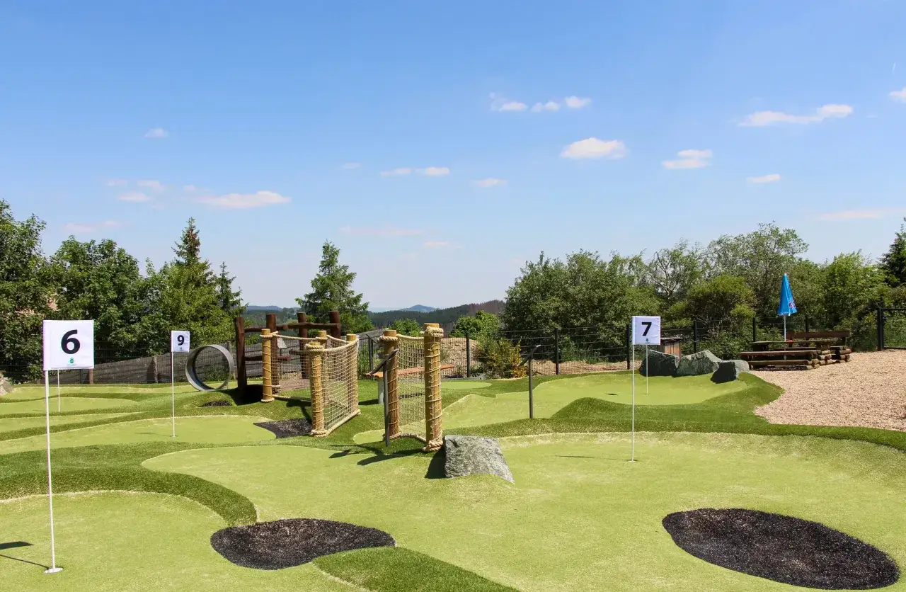 Abenteuer Minigolf NRW: Grüne Bahnen mit Hindernissen wie Seilnetzen und einem Reifen unter blauem Himmel.