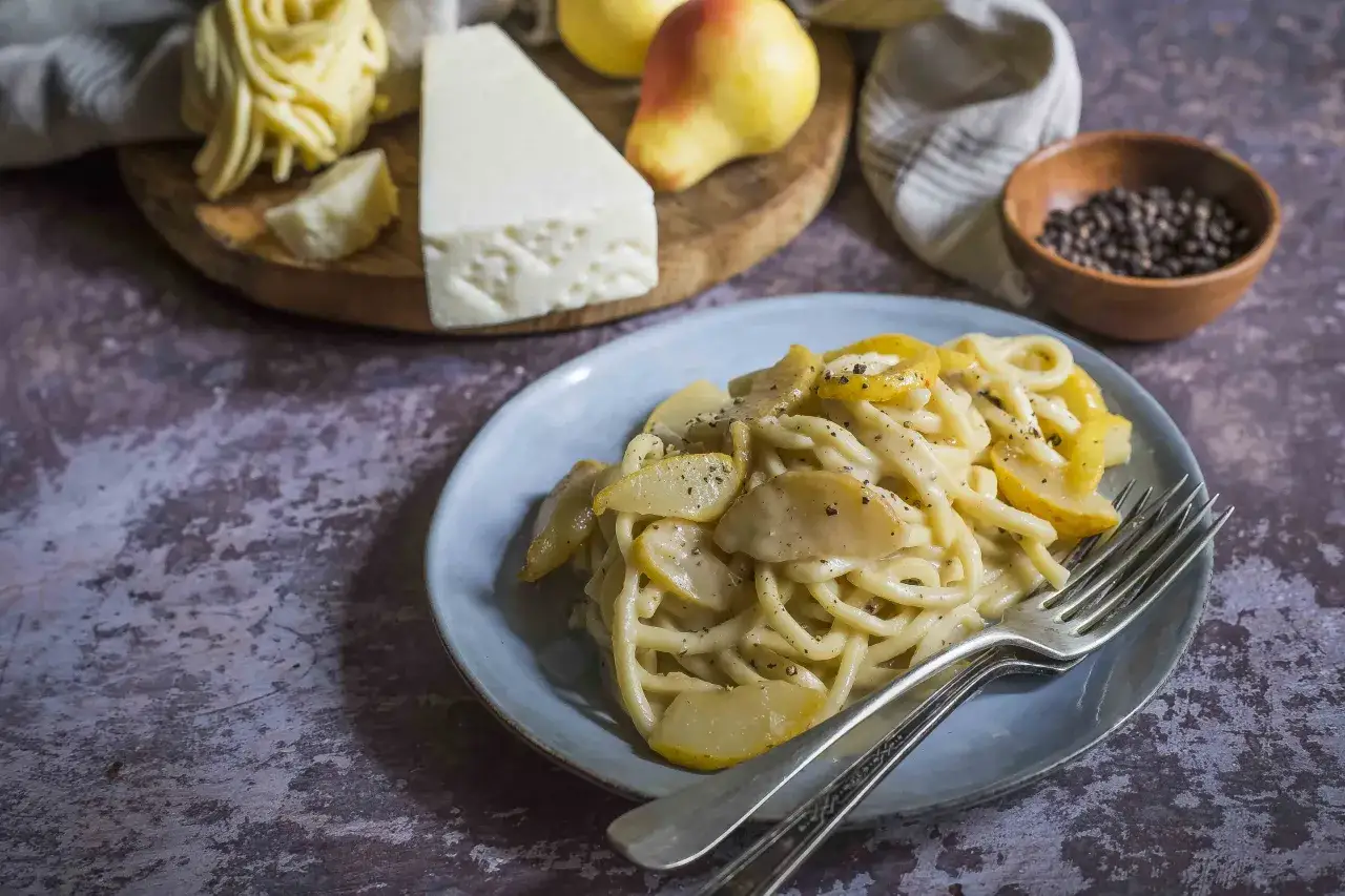 Un piatto di spaghetti conditi con una cremosa salsa al formaggio e pezzetti di pera. Sullo sfondo, ingredienti freschi: formaggio, pere e pepe nero.