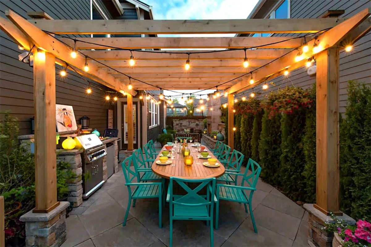 Ideas para iluminar una p&eacute;rgola: luces de cadena crean un ambiente c&aacute;lido sobre una mesa de comedor exterior y parrilla.