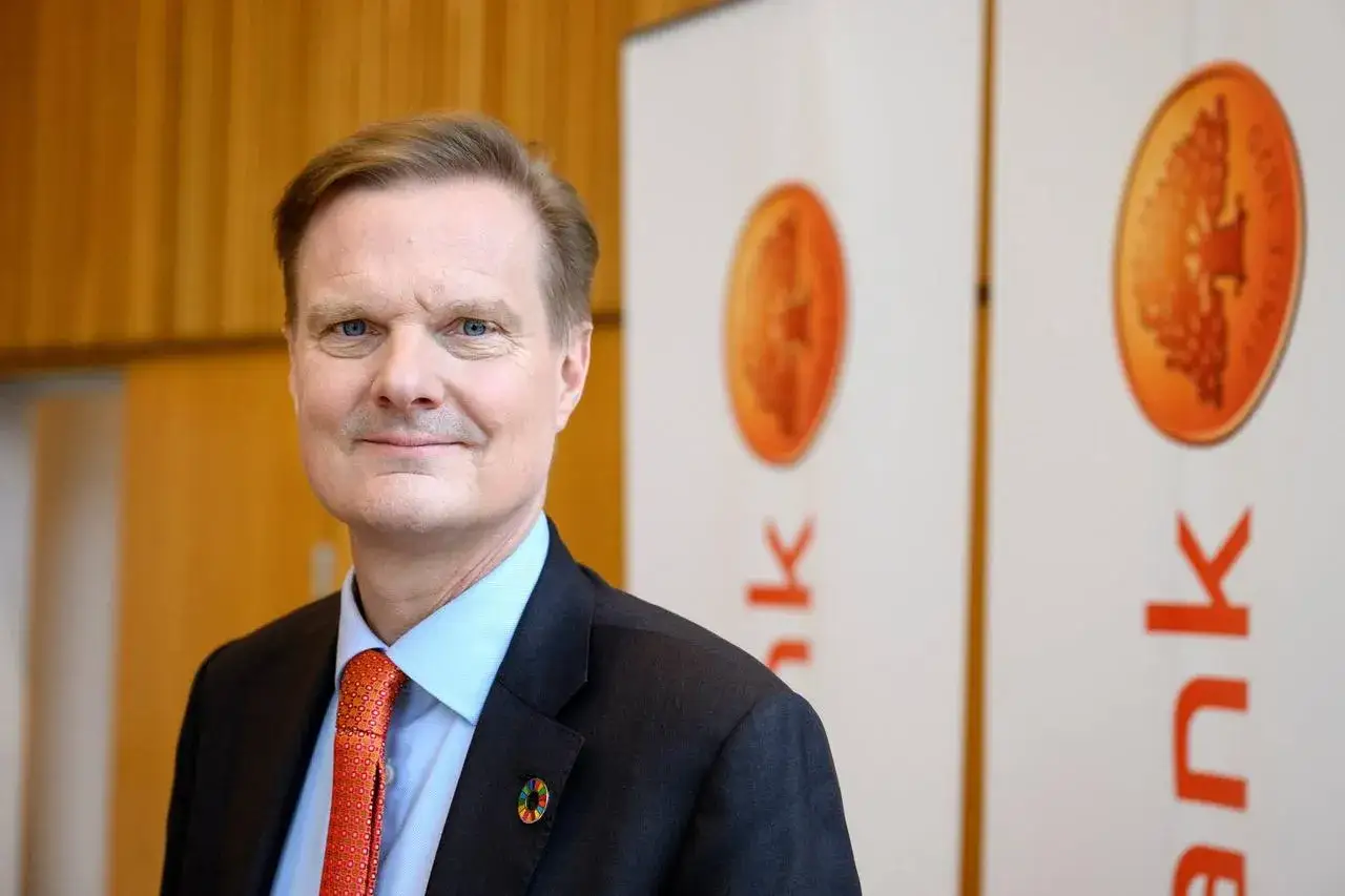 Swedbank VD med bl&aring; &ouml;gon och orange slips, st&aring;r framf&ouml;r Swedbanks logotyp.