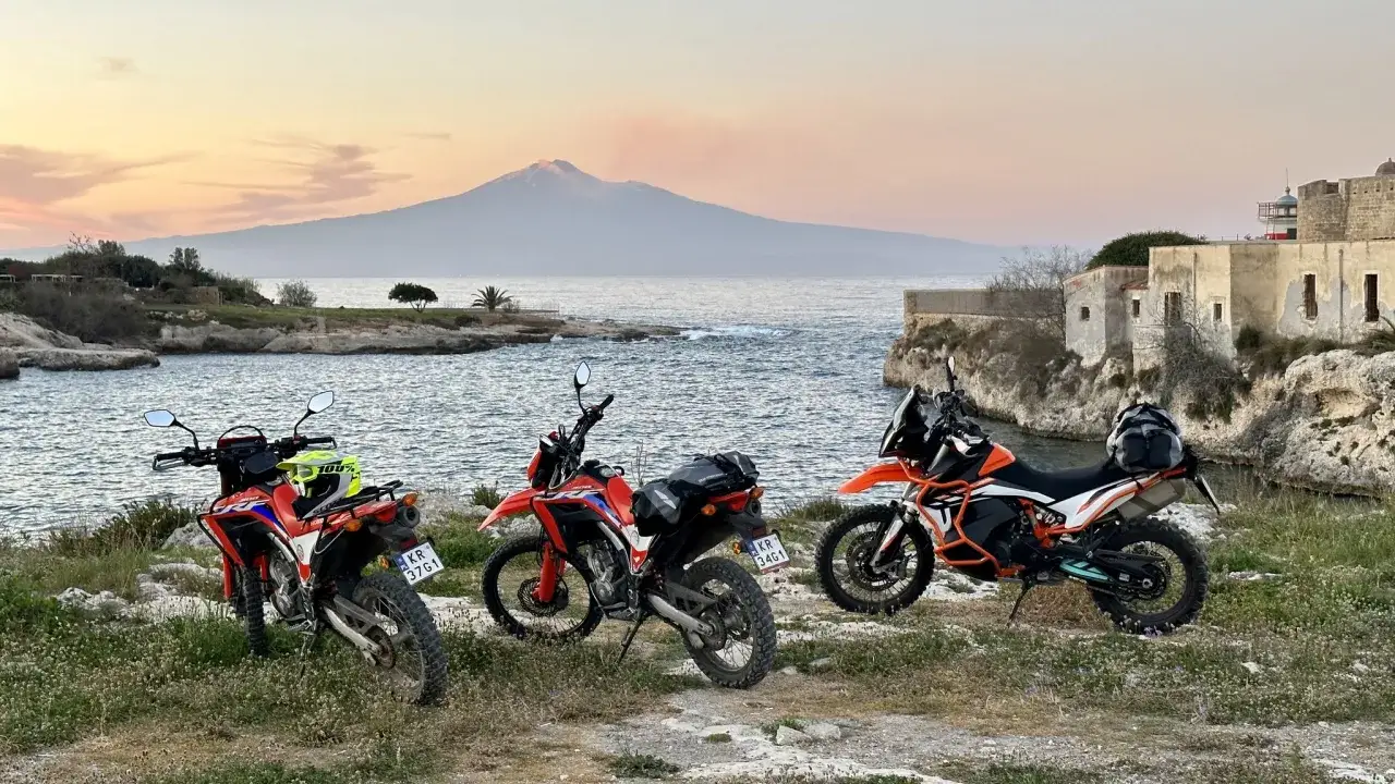 Trzy motocykle terenowe przy morzu, z widokiem na wulkan. Przygotowani na podr&oacute;ż z Livorno do Palermo.
