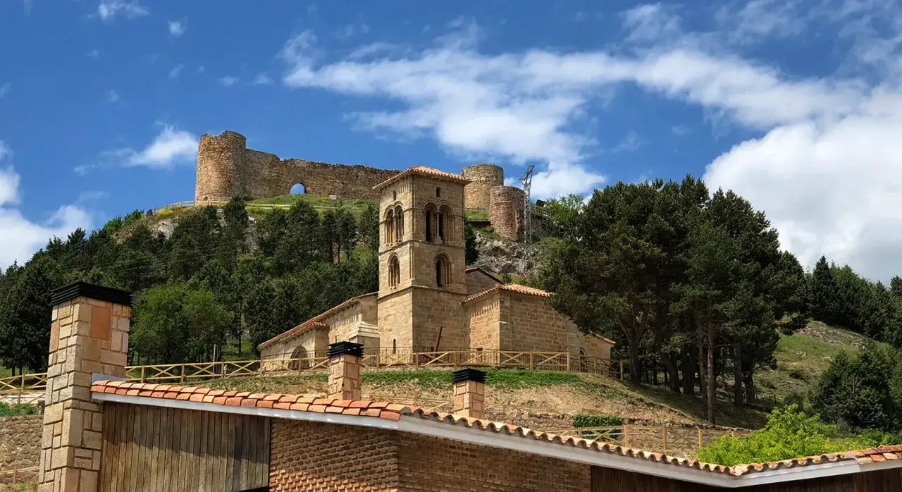Iglesia románica y castillo en ruinas en una colina verde. Un lugar histórico que ver en Palencia, con cielos azules y nubes.
