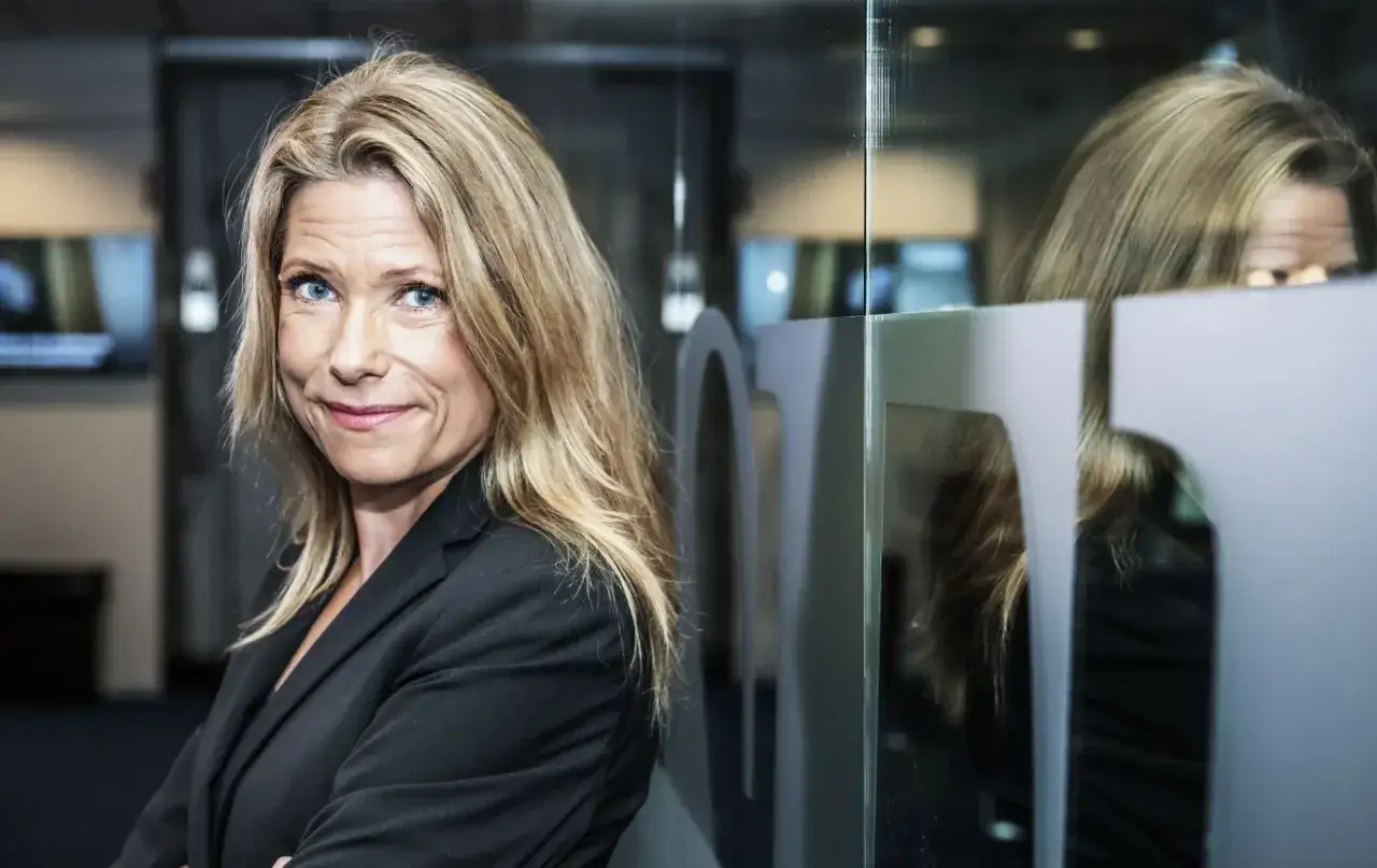 Petra Nordlund, med sitt blonda hår och blå ögon, ler mot kameran. Hon bär en svart kavaj och står framför en glasvägg med en spegelbild.