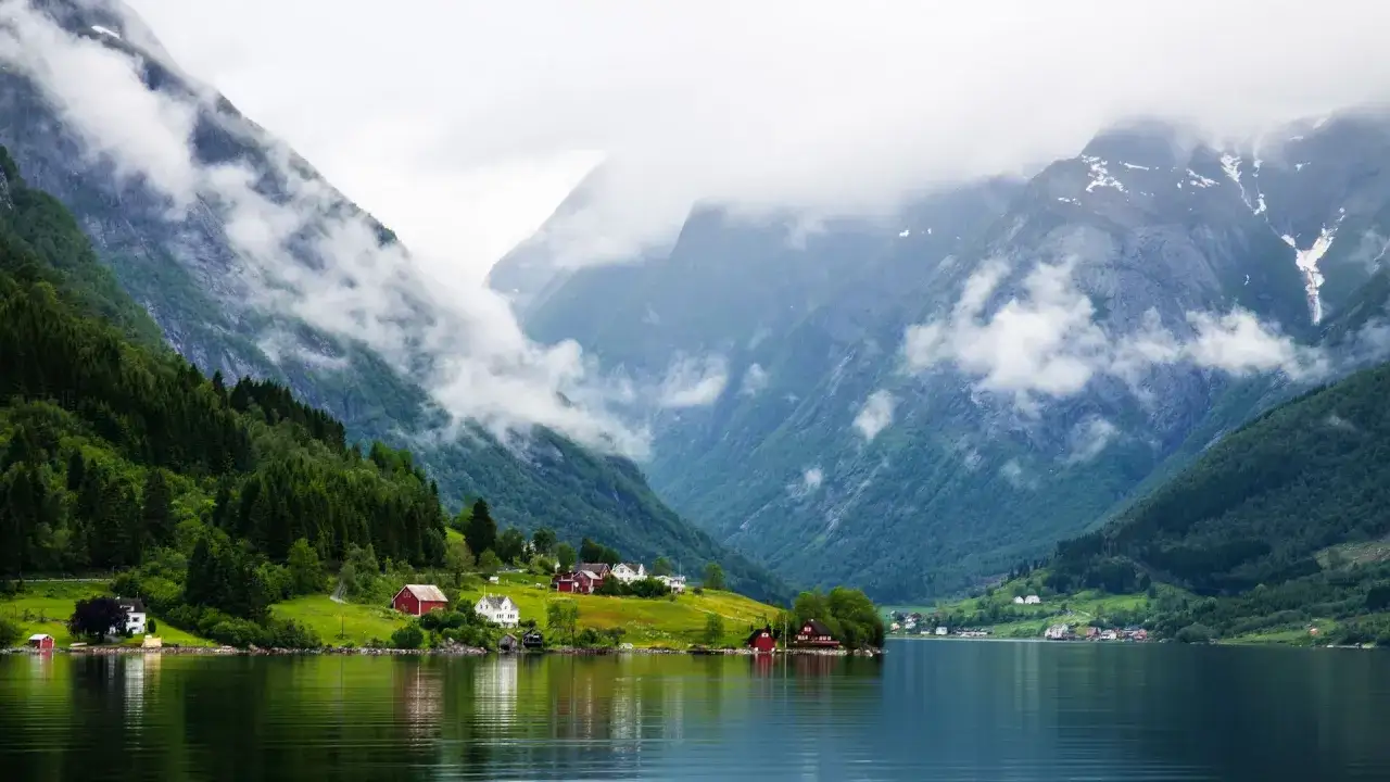 Paysage de fjord brumeux en Norvège, avec des montagnes verdoyantes, des maisons rouges et blanches sur la rive, et l'eau calme reflétant le ciel couvert.