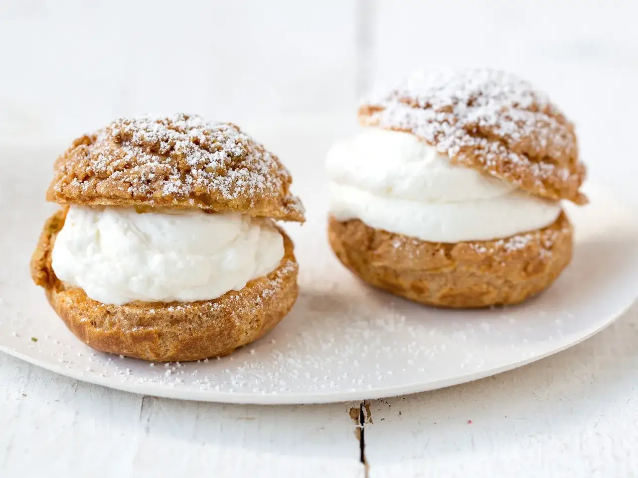 Deux choux à la crème généreusement garnis de crème fouettée, saupoudrés de sucre glace sur une assiette blanche.
