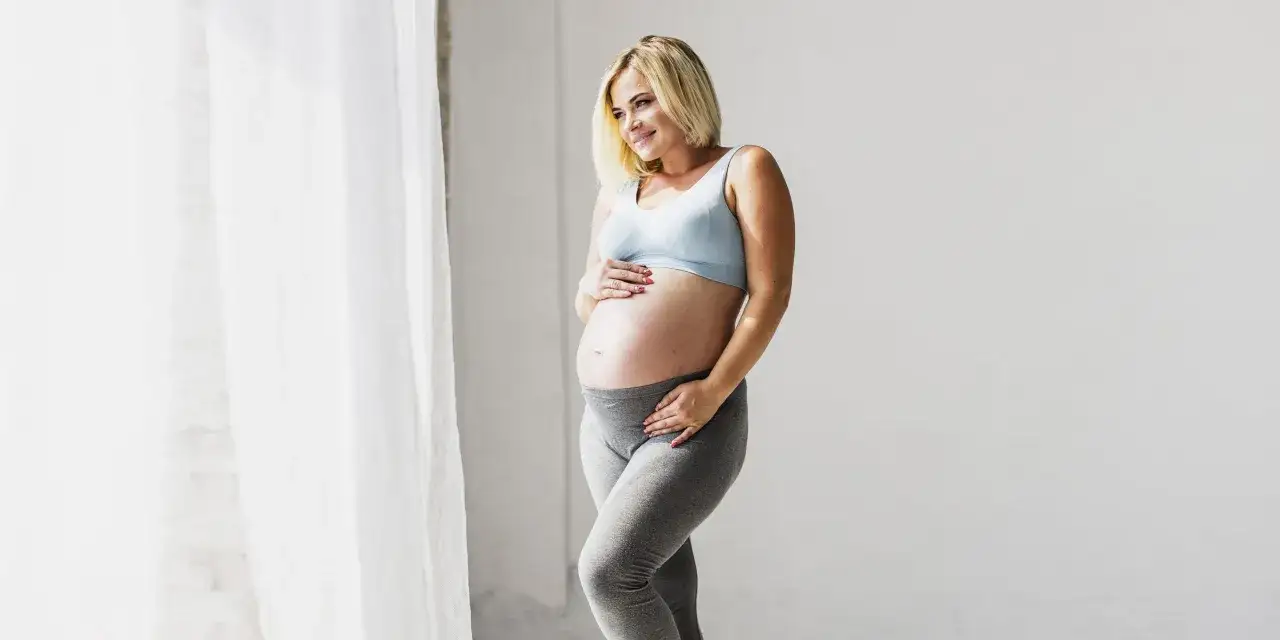 Uśmiechnięta ciężarna kobieta w sportowym topie i legginsach, trzymająca się za brzuch. Dyskutuje o wysokim cholesterolu w ciąży.