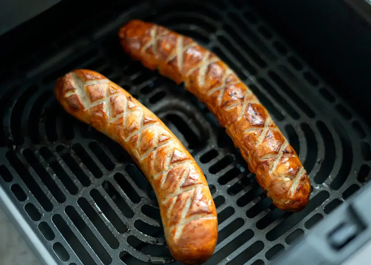 Zwei goldbraune Bratw&uuml;rste mit Rautenmuster liegen auf dem Rost eines Airfryers und werden knusprig.