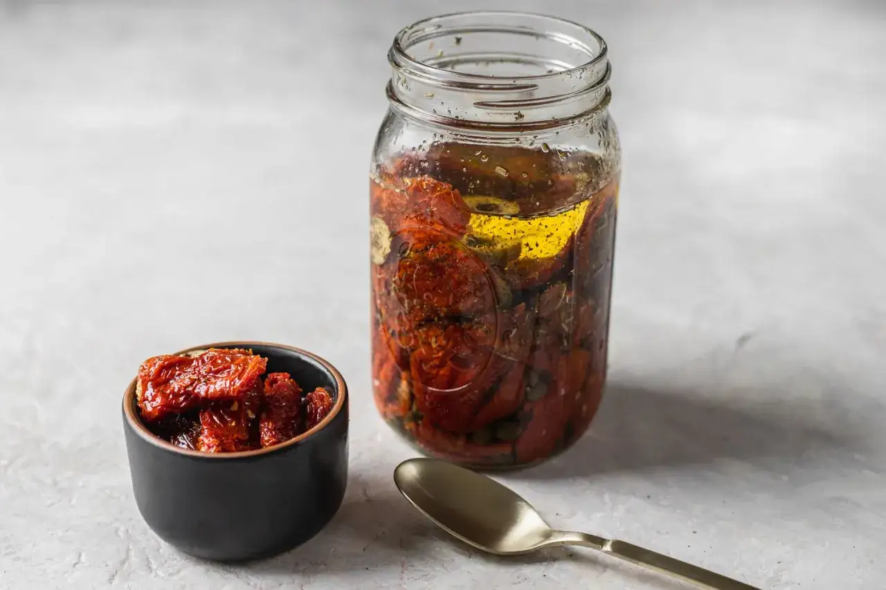Un tarro de cristal lleno de tomate seco en aceite, junto a un cuenco con más tomates y una cuchara.