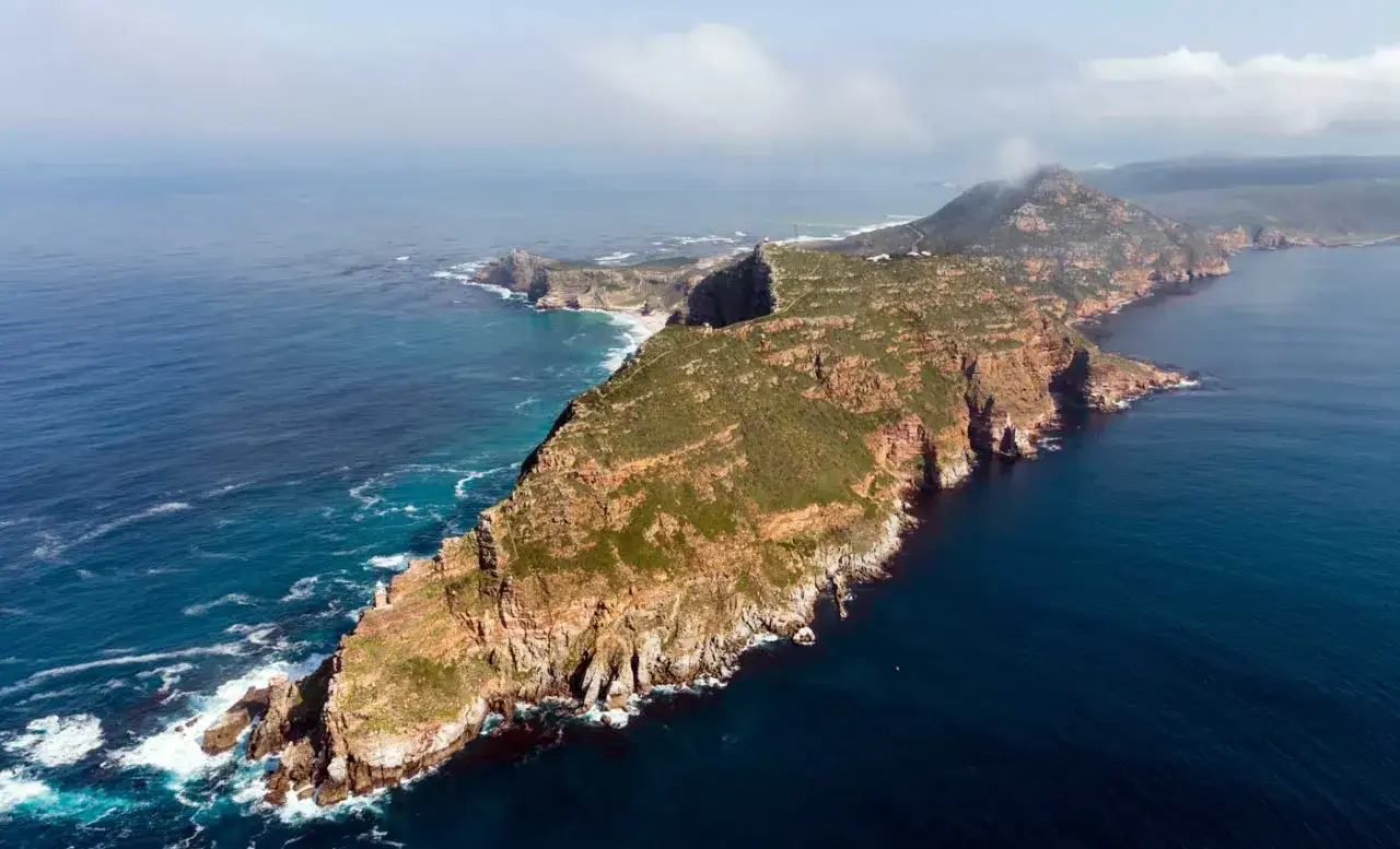 Luftaufnahme der felsigen Kap-Halbinsel mit grüner Vegetation, die ins tiefblaue Meer abfällt. Wellen schlagen an die Küste.