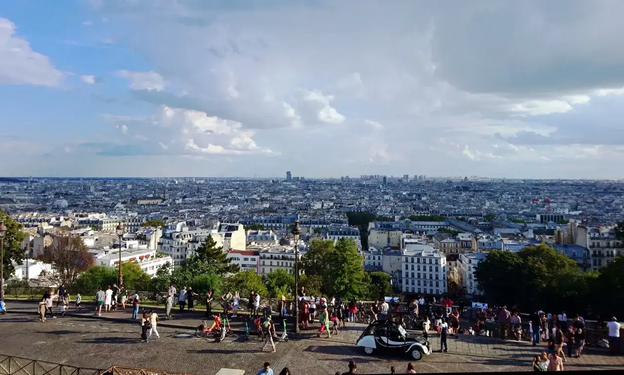 Widok na Paryż z wzgórza Montmartre. Tłum ludzi spaceruje po brukowanym placu, podziwiając panoramę miasta.