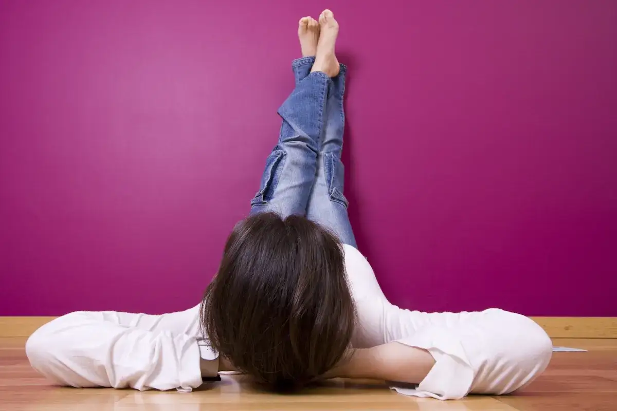 Frau mit hochgelegten Beinen an einer lila Wand. Ein entspannter Moment, der Linderung f&uuml;r schwere Beine verspricht, vielleicht mit Hausmitteln.