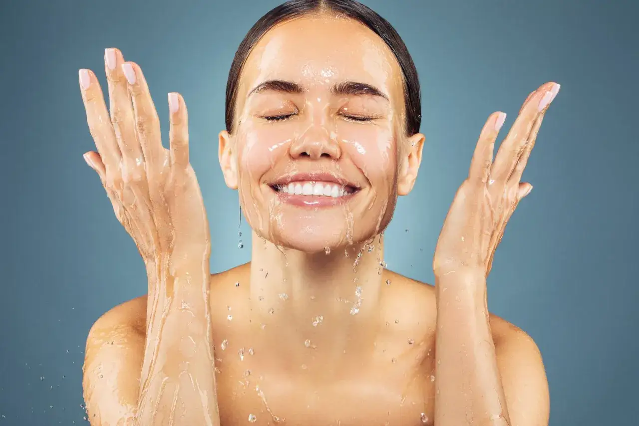 A woman smiles with her eyes closed, splashing water on her face. Her hands are raised, and water cascades down her arms, suggesting a refreshing facial ice bath.