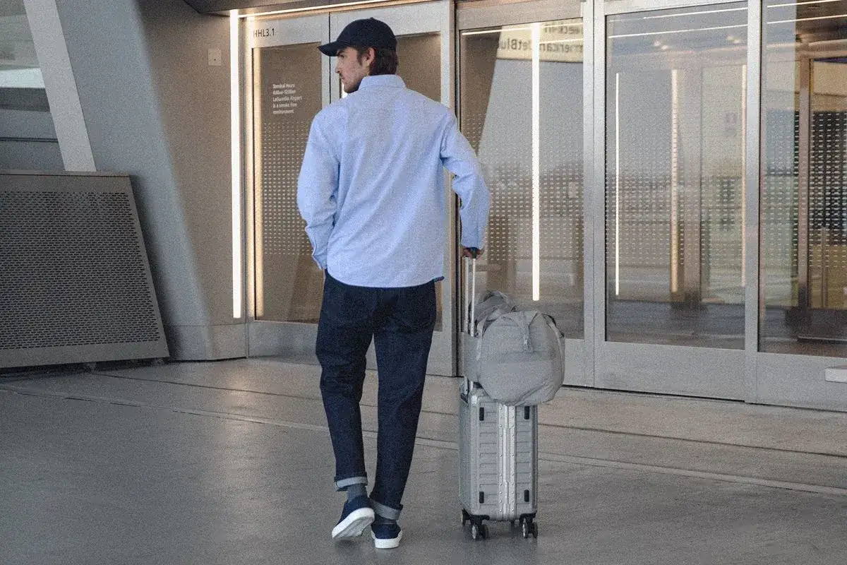 Un homme en jean et chemise bleue, casquette, avec une valise et un sac de voyage, se pose la question : quelles chaussures porter avec un jean homme pour un look décontracté et stylé ?