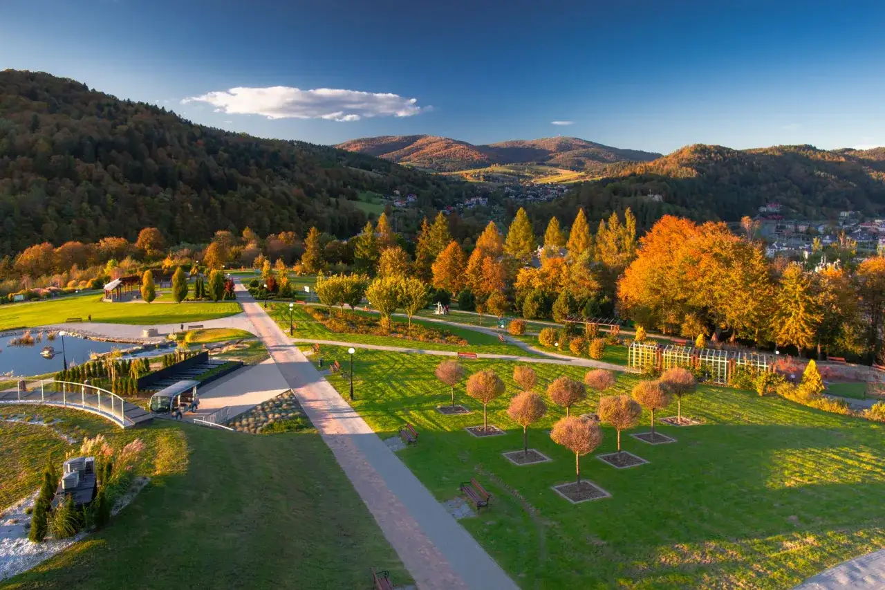 Jesienny park z alejkami, stawem i drzewami w barwach **velo natura**. W tle góry i miasteczko.
