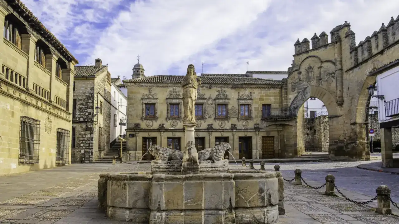 Plaza con fuente de leones y figura central, rodeada de edificios históricos y un arco monumental en Ubeda y Baeza.