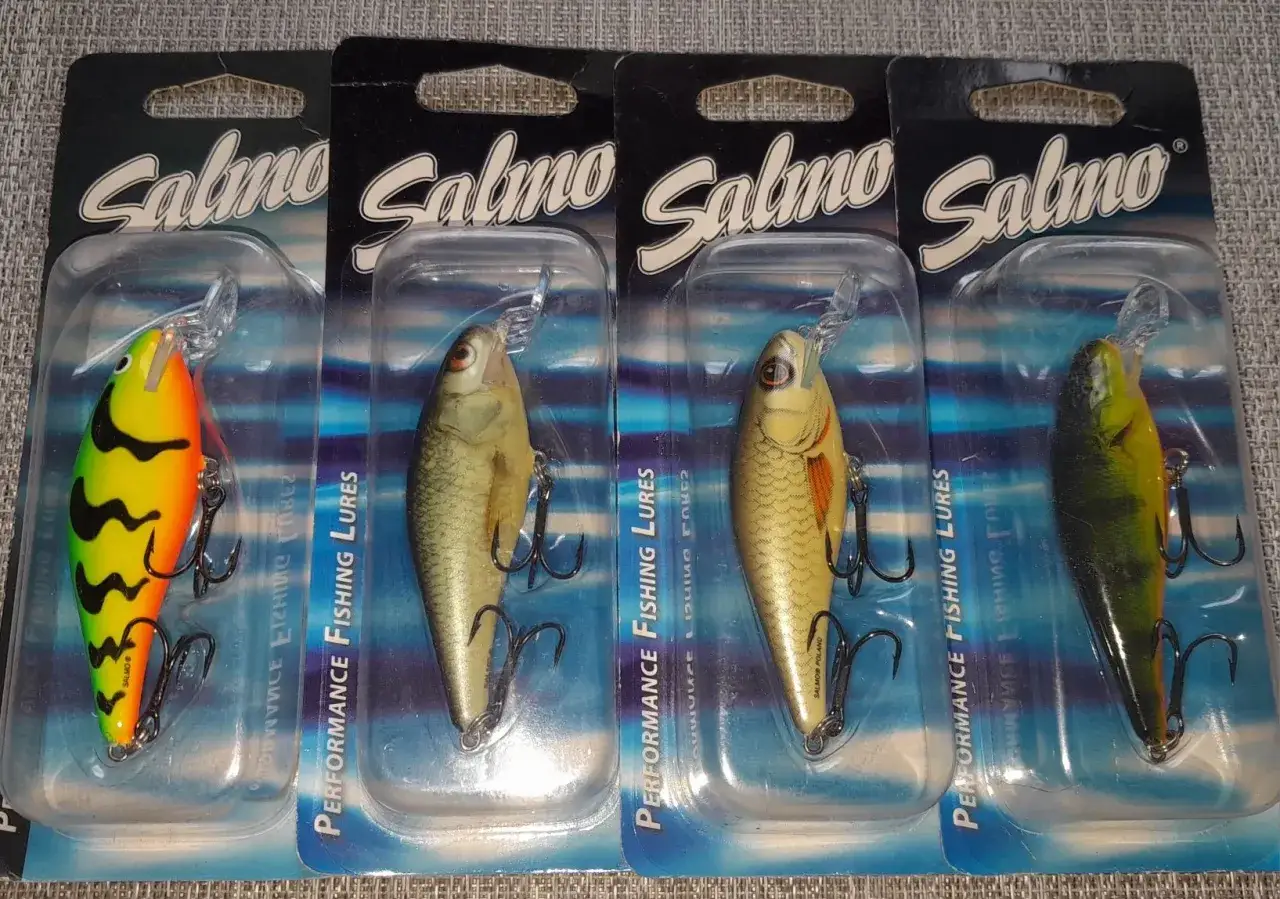Cztery przynęty Salmo w opakowaniach. Jedna jaskrawożółta z czarnymi falami, pozostałe imitują ryby. Gotowe na łowisko, niczym salmo executor.