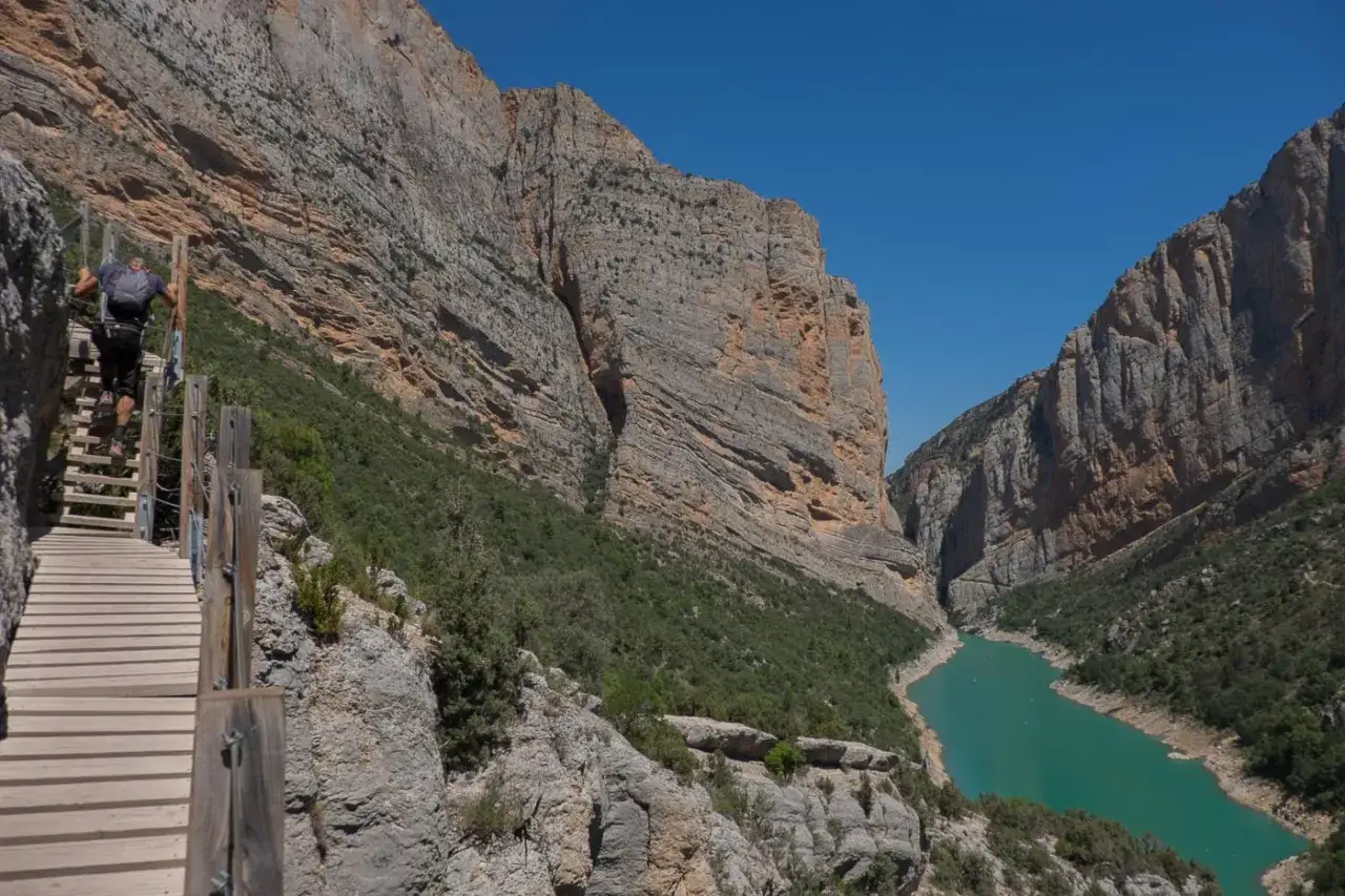 Un randonneur escalade les **pasarelas de Montfalcó**, un sentier vertigineux au-dessus d'un canyon aux falaises imposantes et d'une eau turquoise.
