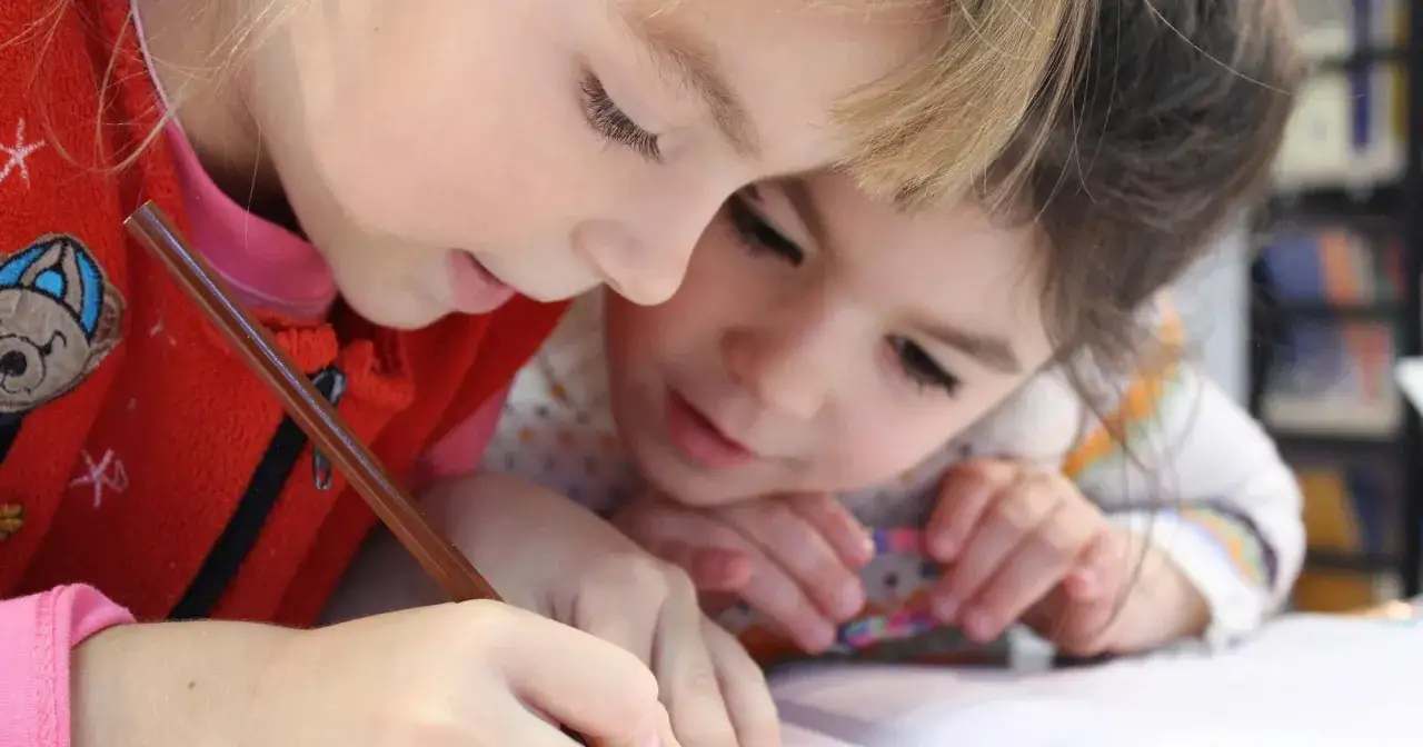Dwie dziewczynki pochylone nad zeszytem, jedna pisze. Zastanawiają się, ile wynosi etat pedagoga szkolnego, by móc mu pomóc.