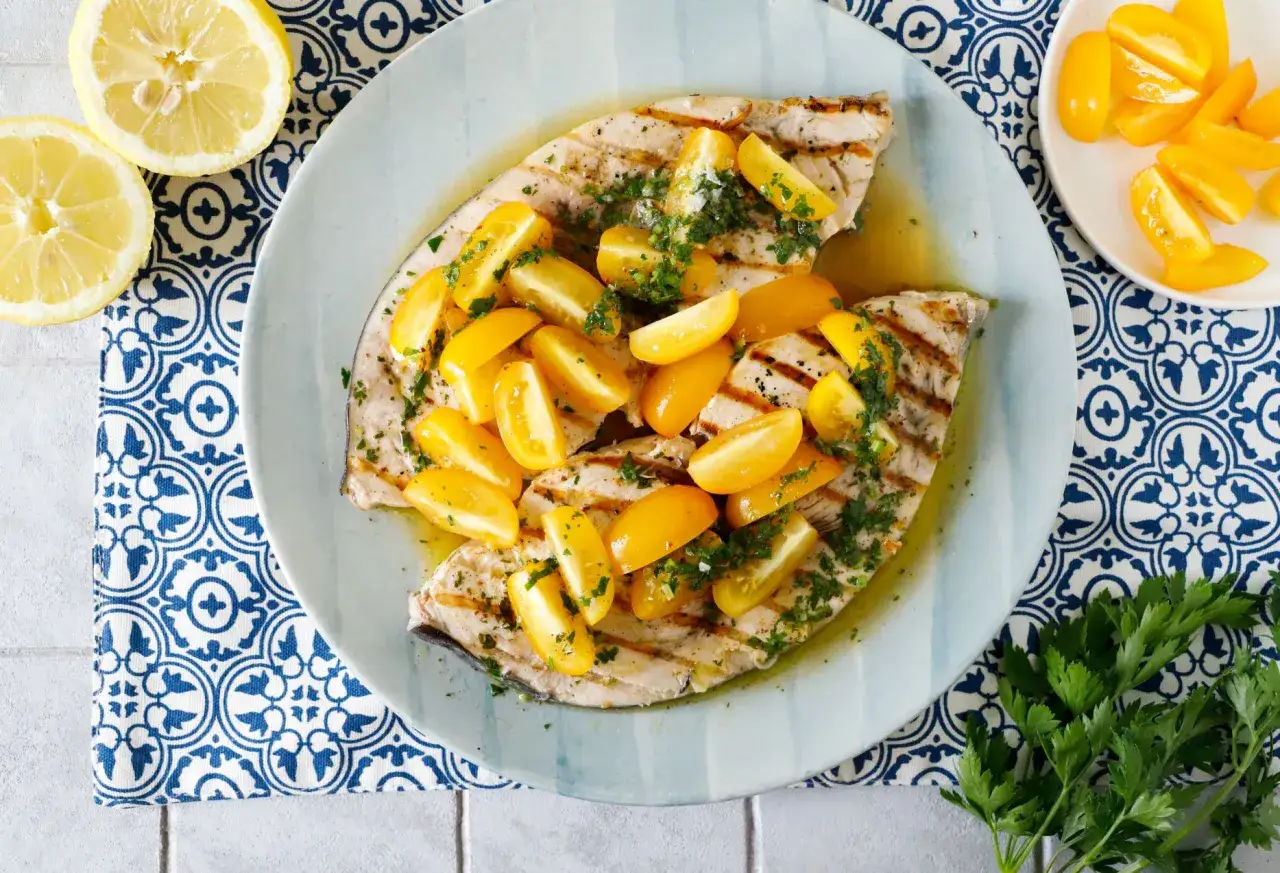 Delizioso pesce spada alla griglia, guarnito con pomodorini gialli e prezzemolo fresco, pronto per essere gustato con spicchi di limone.