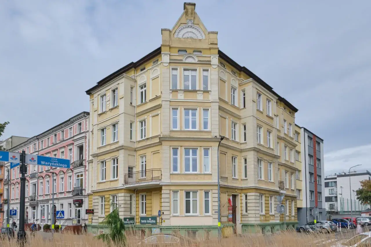 Żółta kamienica z białymi oknami i balkonem. Występ ściany podkreśla architekturę budynku.