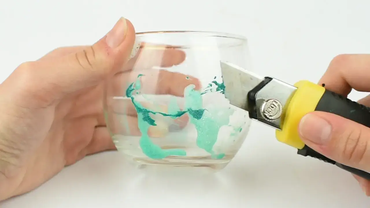 Maintenant, on va enlever peinture sur verre avec un cutter. Une main tient le verre, l'autre manie l'outil pour gratter la peinture verte.