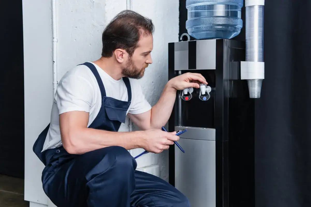 Un technicien en bleu de travail effectue l'entretien d'une fontaine &agrave; eau, v&eacute;rifiant les robinets chauds et froids.