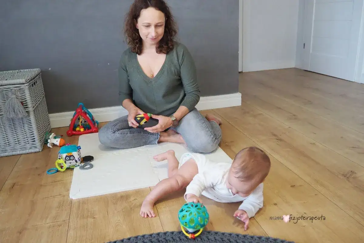 Mama i dziecko bawią się na macie. Dziecko próbuje przyjąć pozycję **siad w literę w**, sięgając po zabawkę.