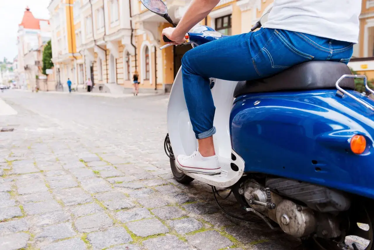 Un uomo in jeans guida uno scooter blu su una strada acciottolata, un'immagine che evoca la libertà e lo spirito dell'art 52 cds.