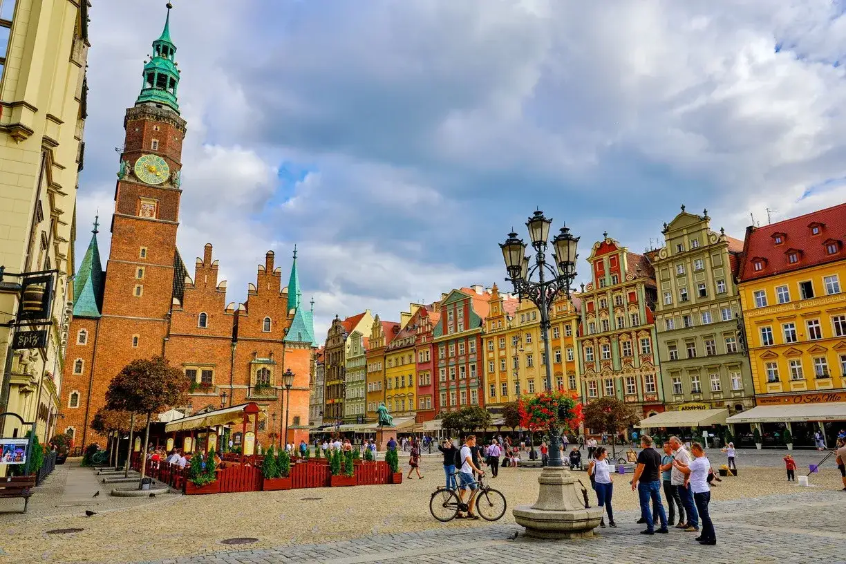 Tętniący życiem rynek Starego Miasta z ratuszem i kolorowymi kamienicami. Ludzie spacerują, siedzą w ogródkach kawiarnianych, a rowerzysta przejeżdża przez brukowany plac.