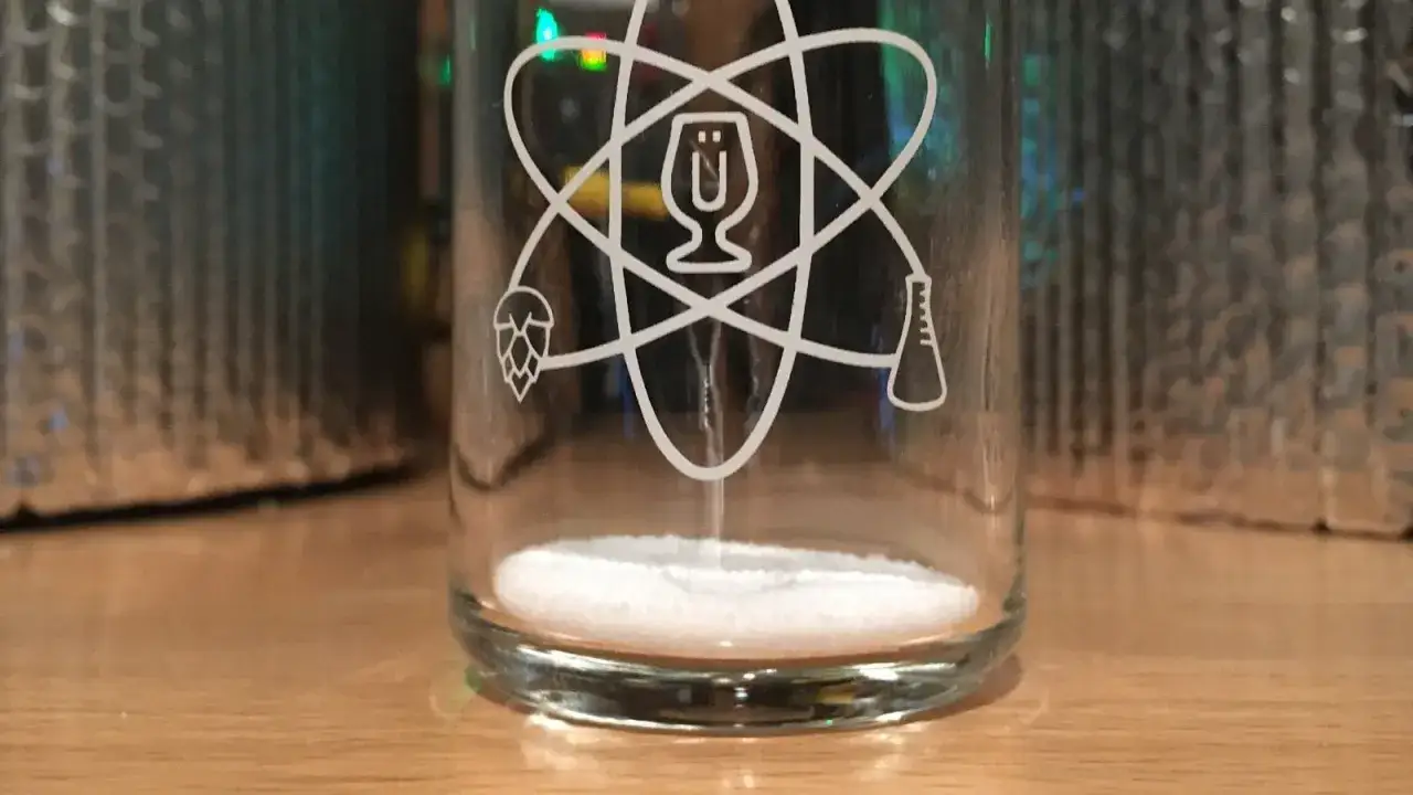 Szklanka z motywem atomu, w której znajduje się biały proszek. Na szkle widnieje chmiel, kieliszek i kolba. Może to być goryczka szorstka.
