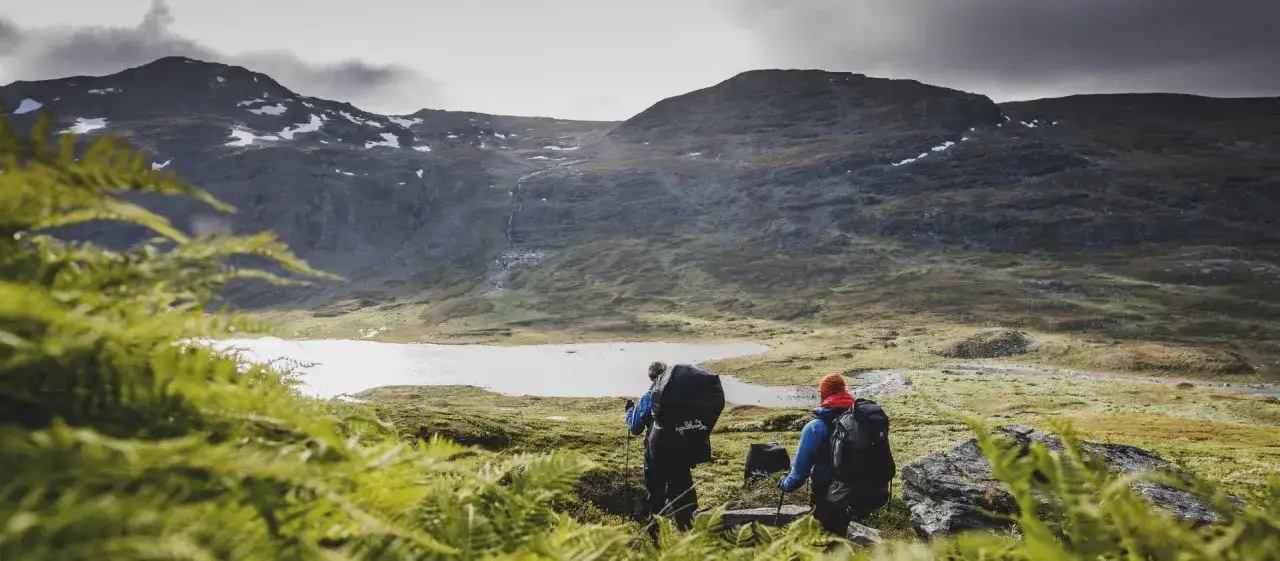 Tv&aring; vandrare med stora ryggs&auml;ckar g&aring;r genom ett bergigt landskap med en sj&ouml; i bakgrunden. En fantastisk vy f&ouml;r vandring i Sverige.