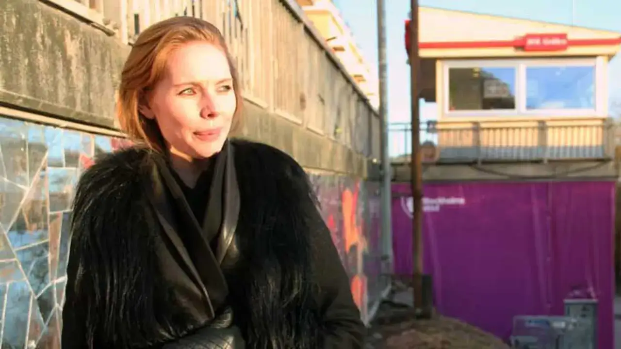 Karin Winther i en svart pälsjacka framför en vägg med mosaik och en lila presenning med texten