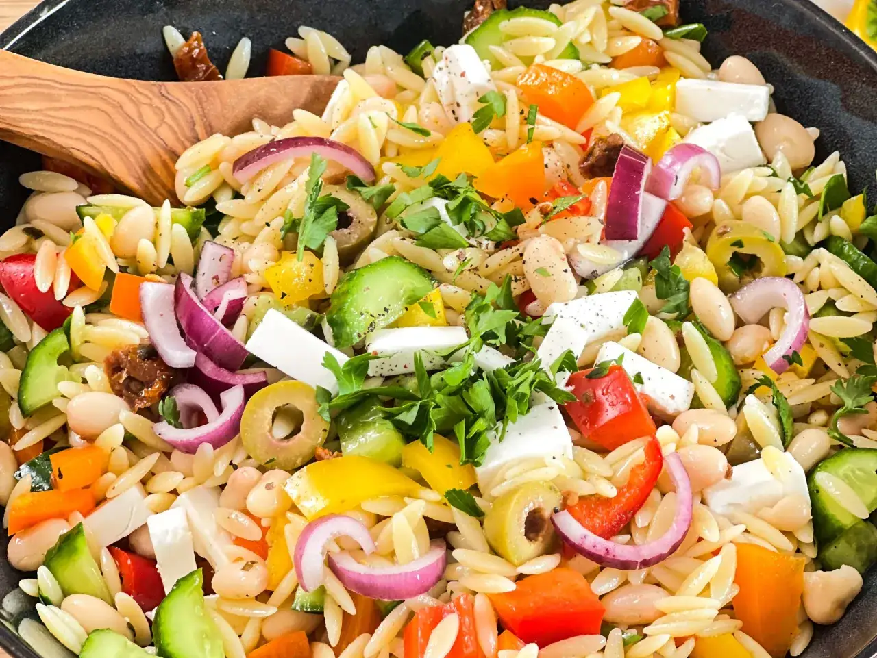 Ein bunter griechischer Nudelsalat mit Orzo-Nudeln, Feta, Oliven, Paprika, Zwiebeln und wei&szlig;en Bohnen, garniert mit frischer Petersilie.