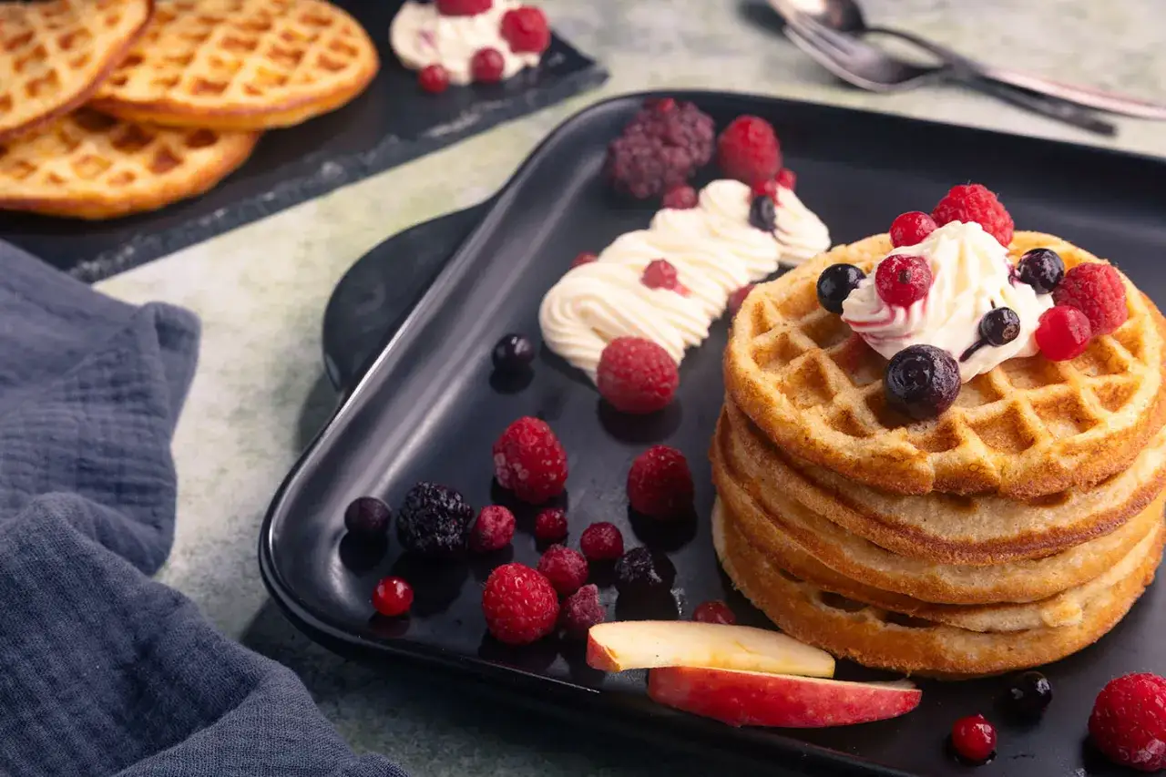 Ein Stapel goldbrauner Waffeln, garniert mit Sahne und Beeren, perfekt f&uuml;r ein Apfelwaffeln Rezept.