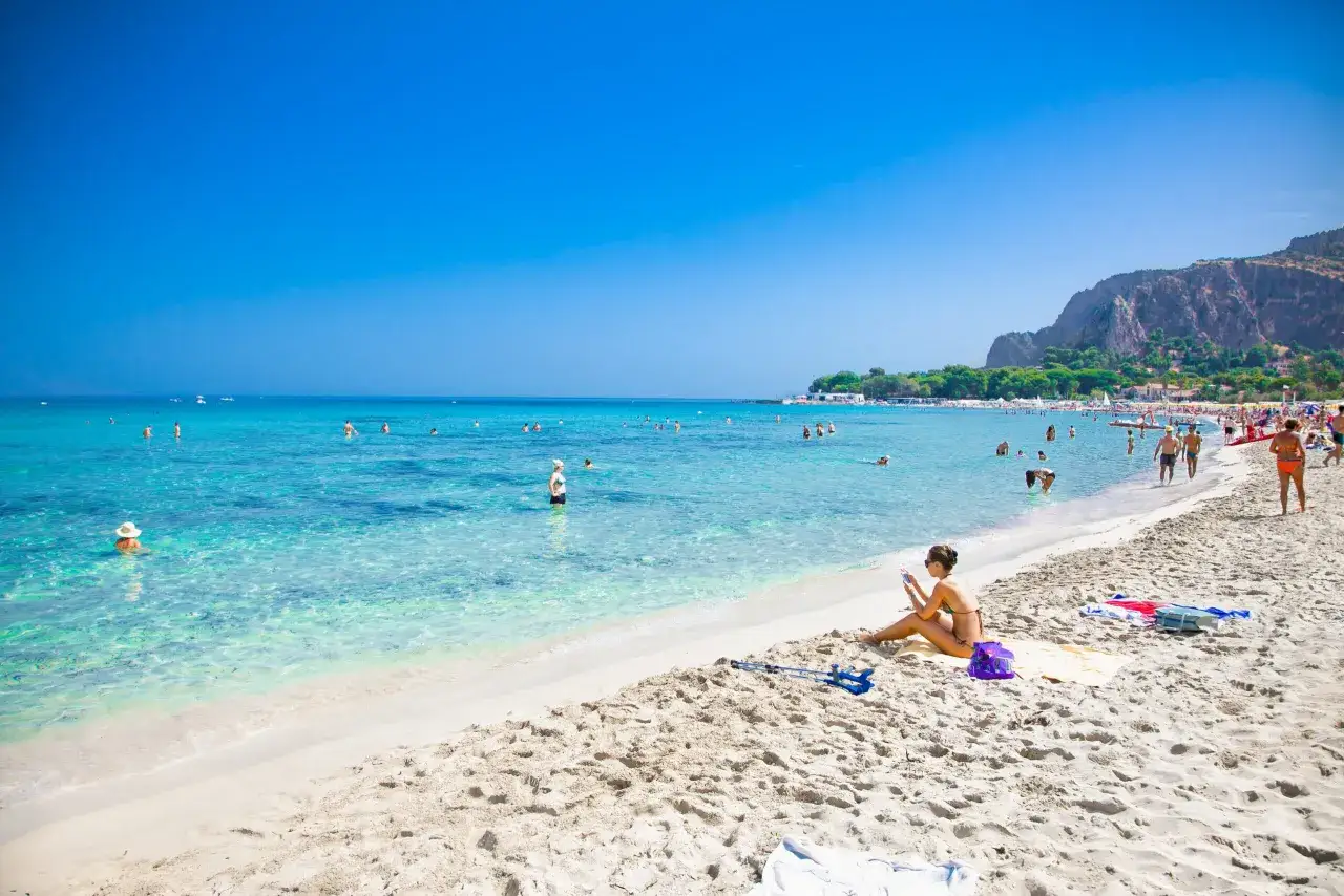 Słoneczna plaża Mondello z turkusową wodą, ludźmi kąpiącymi się i odpoczywającymi na piasku, w tle góry.